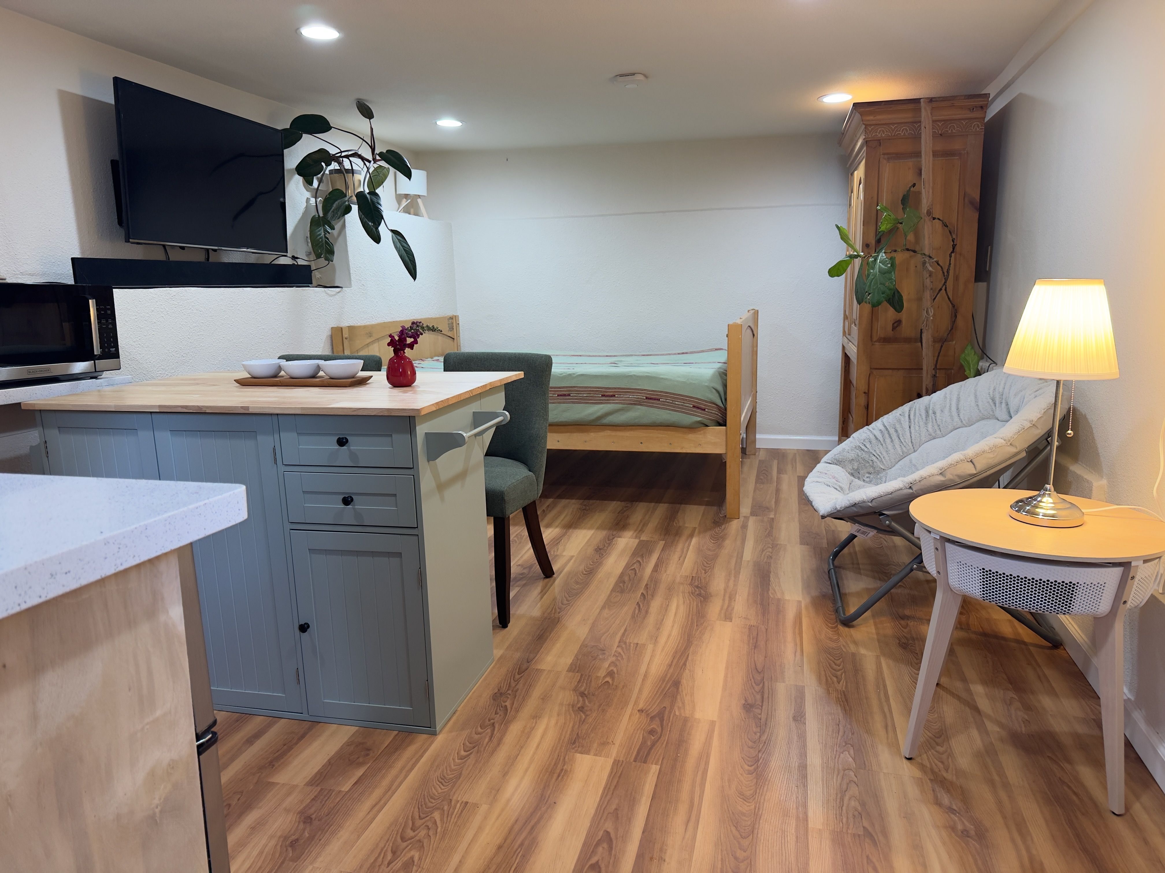 The furnished bedroom suite: an open room with a bed, a kitchenette island with seating, a wall-mounted TV, a wardrobe, and a papasan chair.
