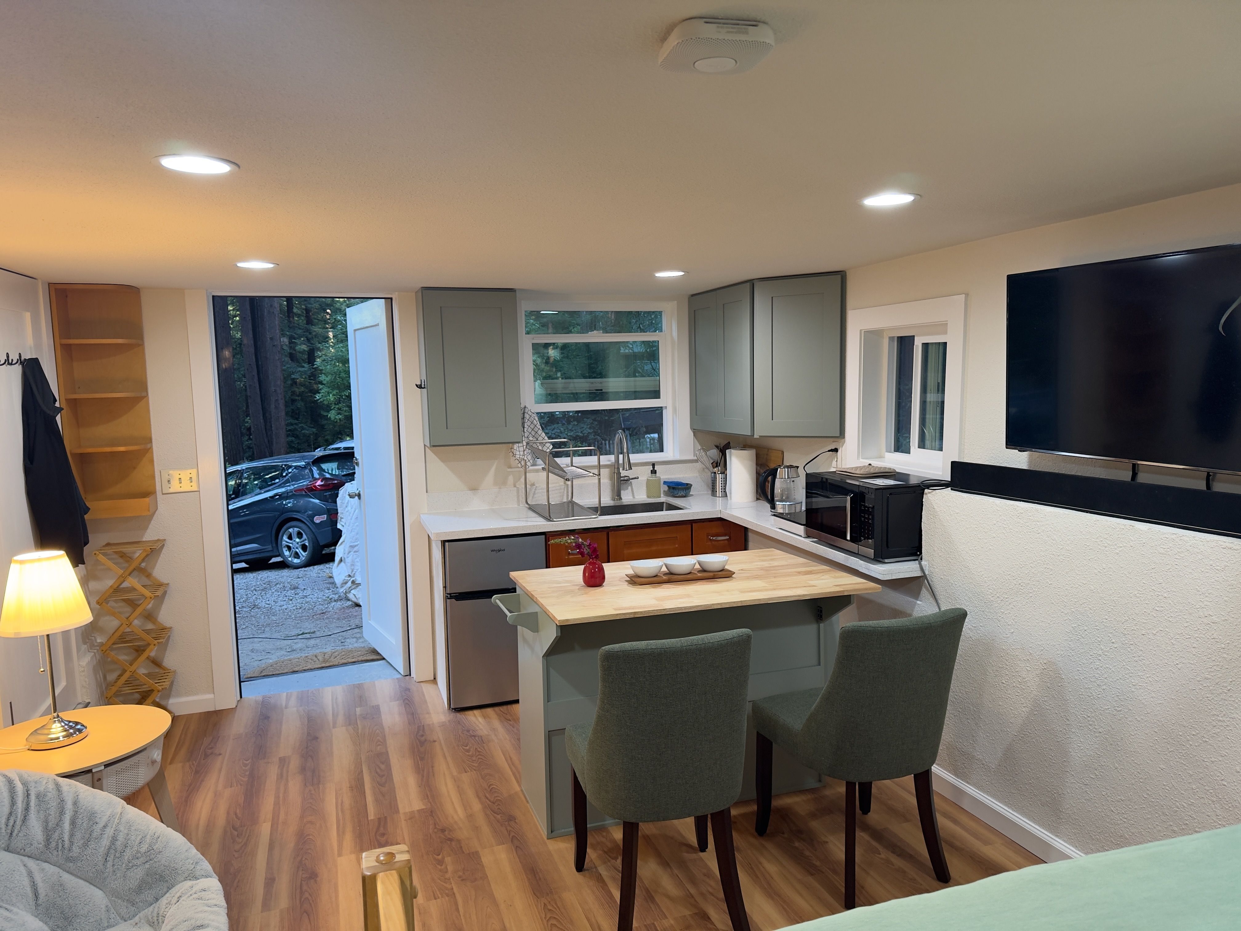 The kitchenette and private entrance: green cabinets, a sink under the window, an induction cooktop, a microwave + air fryer, and a mini-fridge. The exterior door opens to the redwoods and parking. The interior door opens to the bathroom.