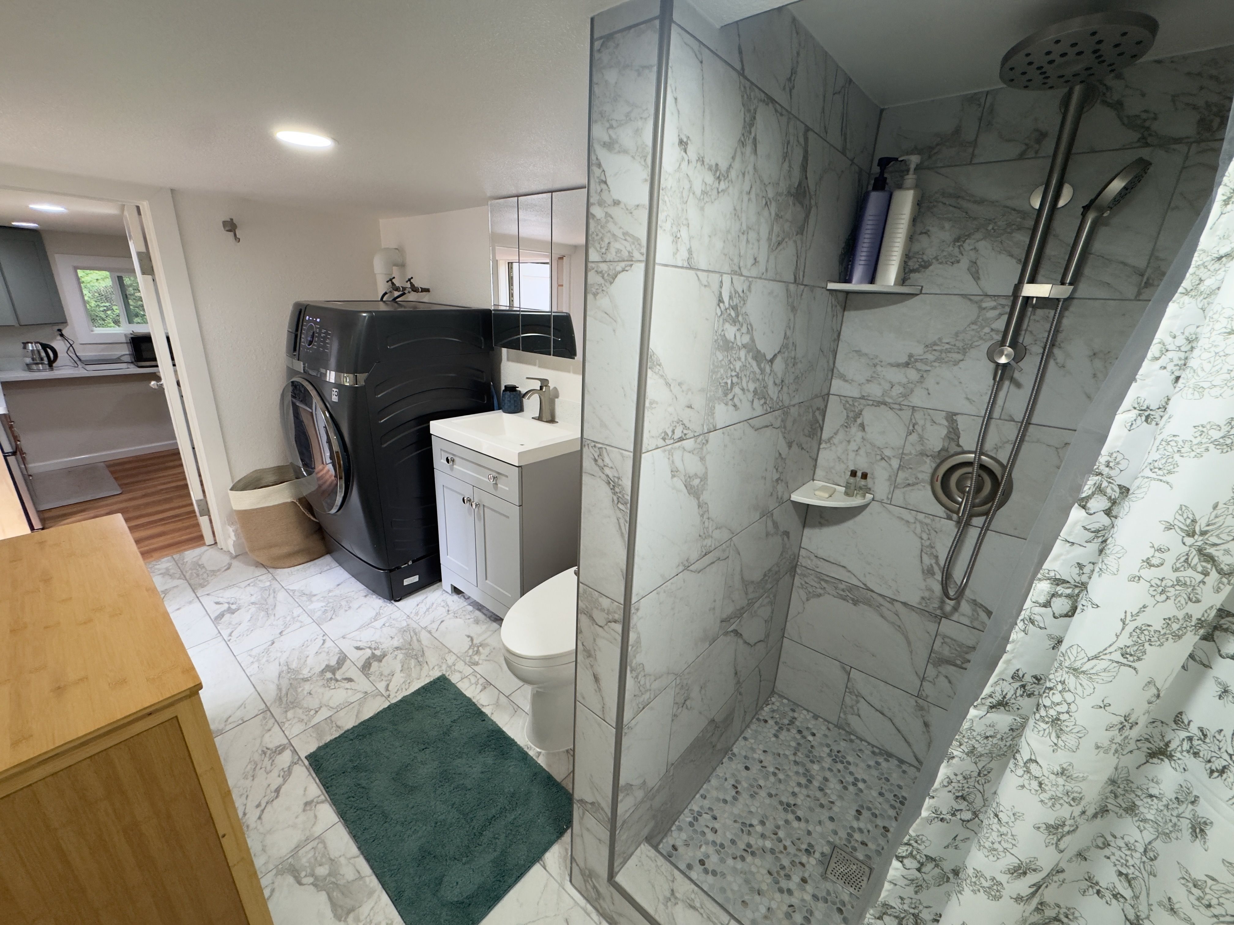 The bathroom from the shower side: a antimicrobial tile walk-in shower, vanity, toilet, and full-size washer+dryer (2.5 hours wash+dry combined), opening onto the bedroom.